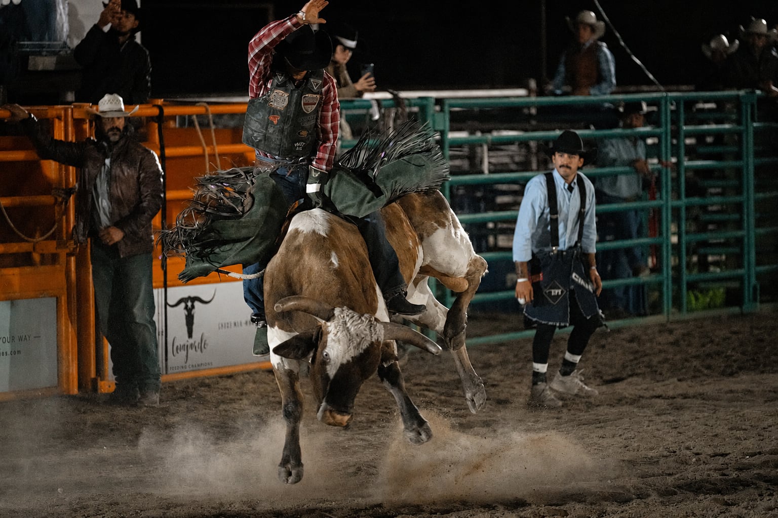 A bull kicks into the air beneath a red-plaid rider as bullfighters watch nearby