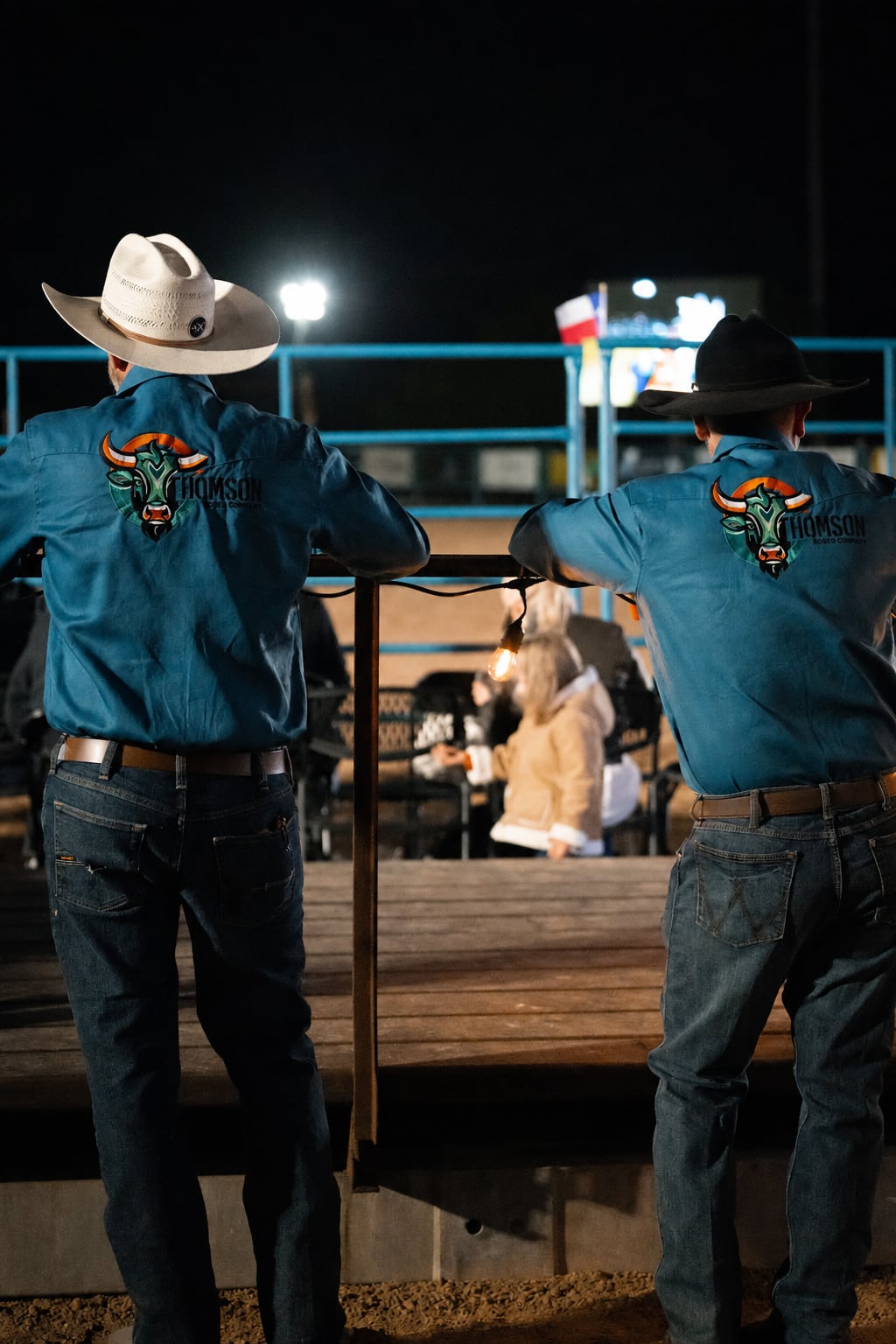 Two A Thomson Rodeo crew members in embroidered teal shirts watch the arena from the rail